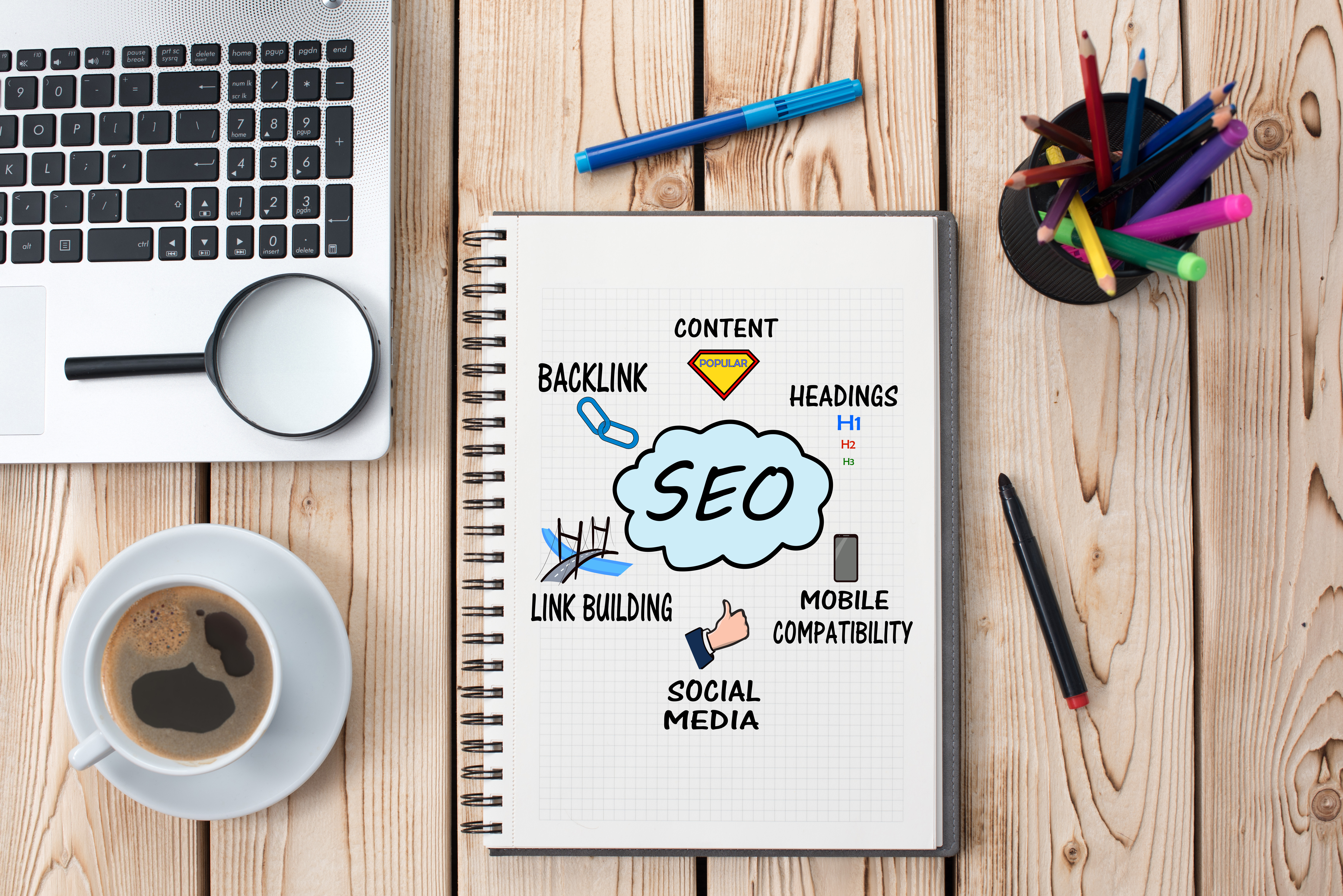 a seo notebook is surrounded by a laptop, cup of coffee, and pens